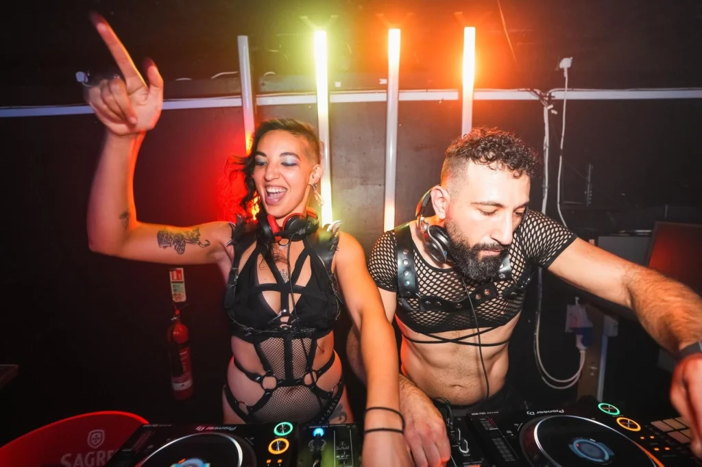Mattana laughing with one hand raised and G Minor focused on the mixer, side by side at the decks under coloured light tubes