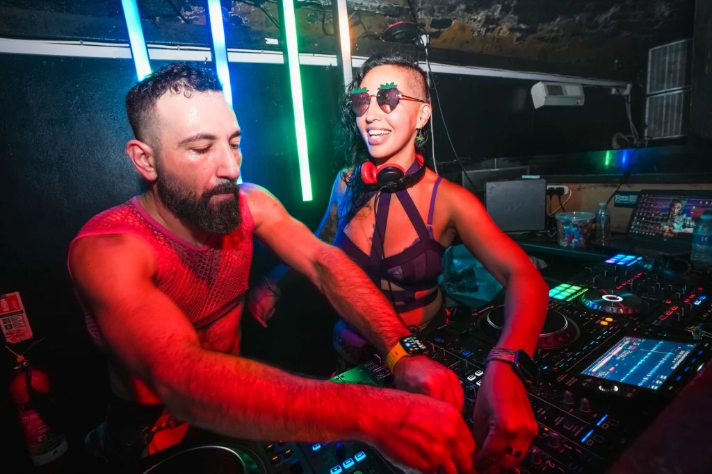G Minor in a red mesh tank and Mattana in heart-shaped sunglasses and red headphones, mixing together on a DJ controller