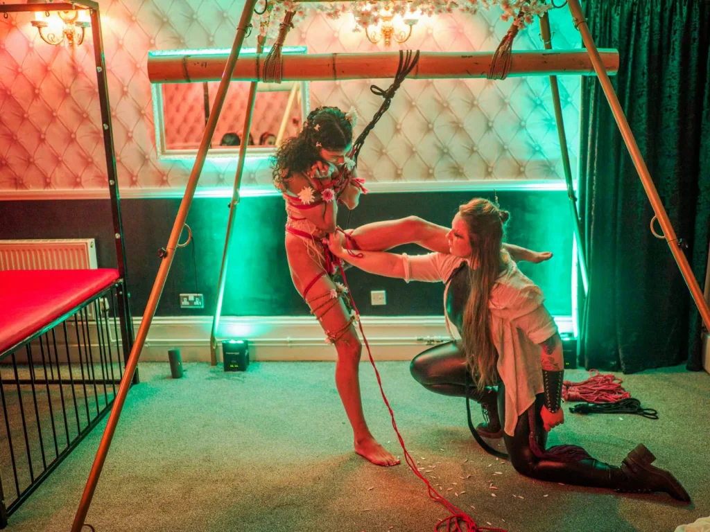 Panthera Valkyrie kneeling beneath the bamboo suspension frame, lifting the bound leg of her rope partner
