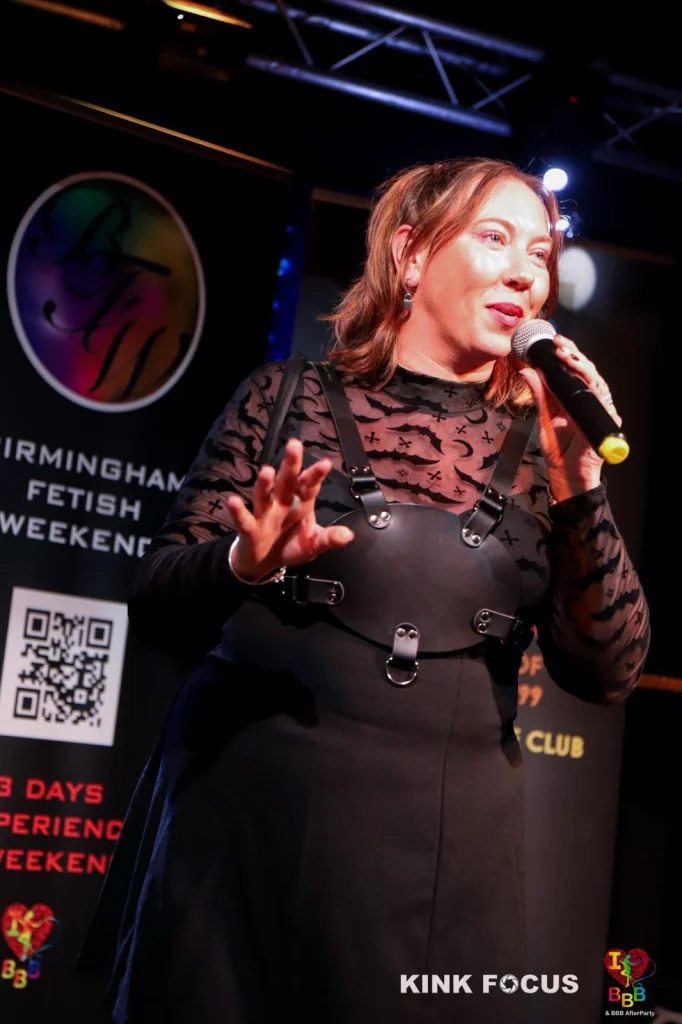 Beastie-FS speaking into a microphone on stage, in a sheer bat-print top and a leather harness