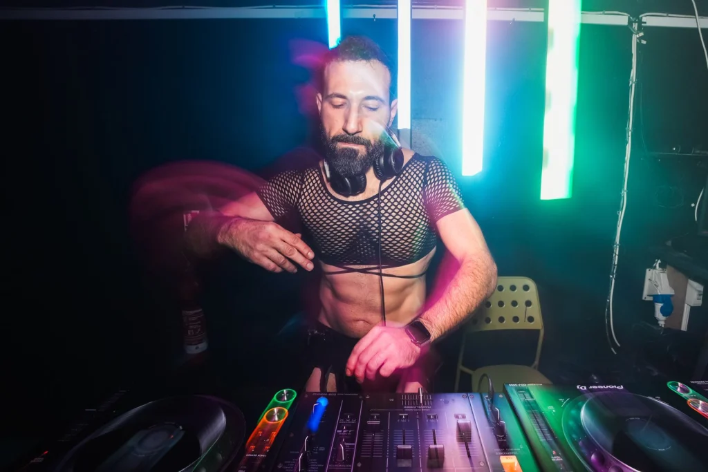 G Minor in a black fishnet crop top with headphones round his neck, at the decks under green and blue light tubes