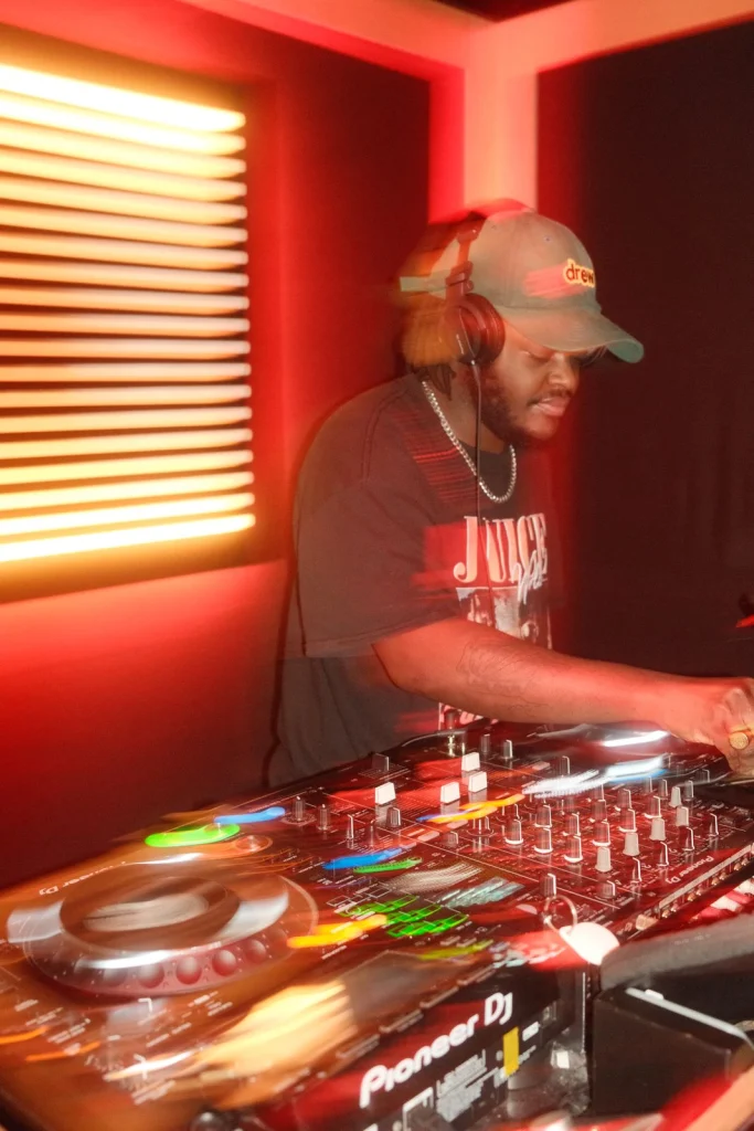 KingPraun in a cap, headphones and a pearl necklace, leaning over the decks in front of a glowing slatted window