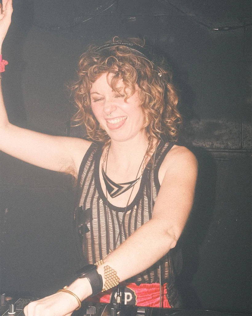 Sian laughing with one arm raised, hands on the decks against a dark wall
